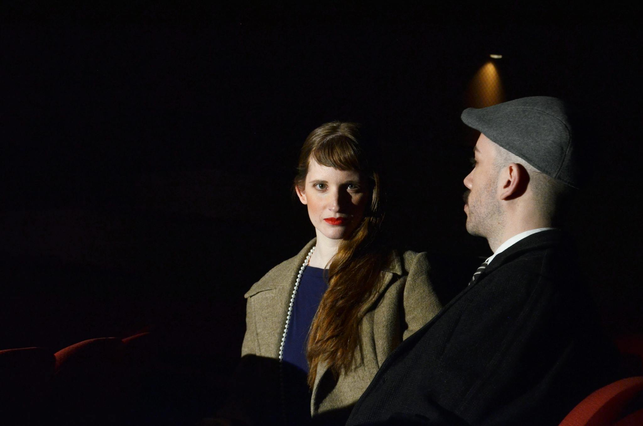 A couple in vintage attire seated in a dimly lit cinema setting, evoking nostalgia.