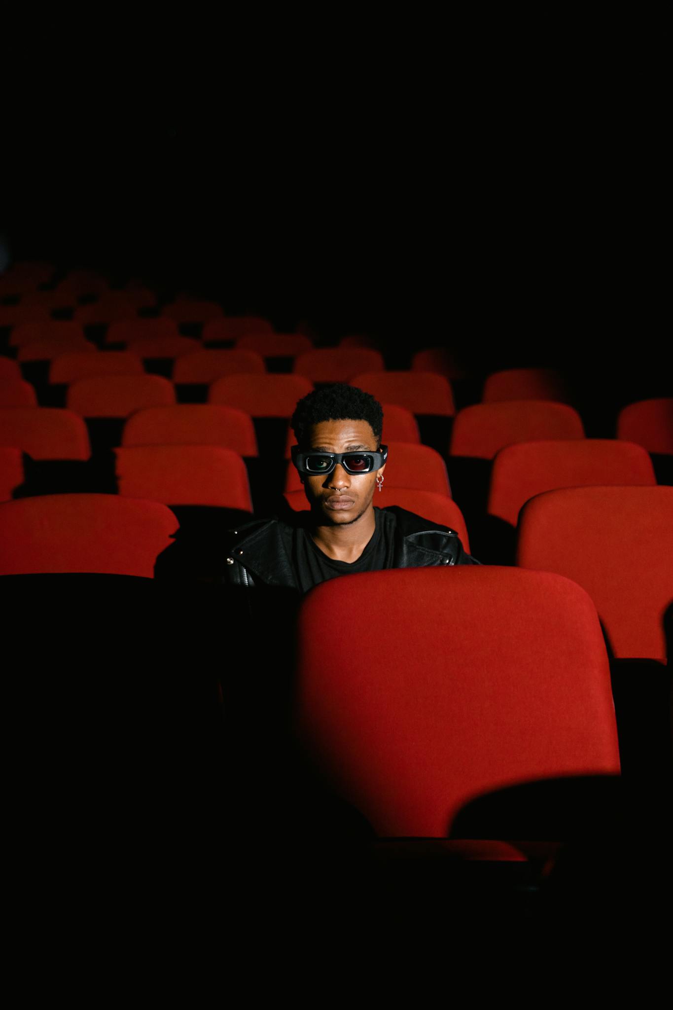 A young man wearing 3D glasses sits alone in a dimly lit, empty cinema.