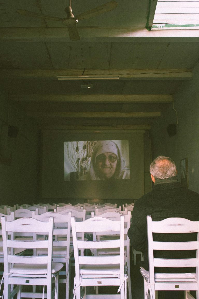 An elderly man sits alone in a theater watching a film screen with white chairs.